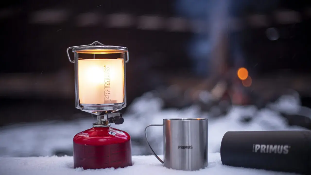En varm primus lampa lyser upp snön bredvid en mugg och en termos. I bakgrunden syns en lägereld.