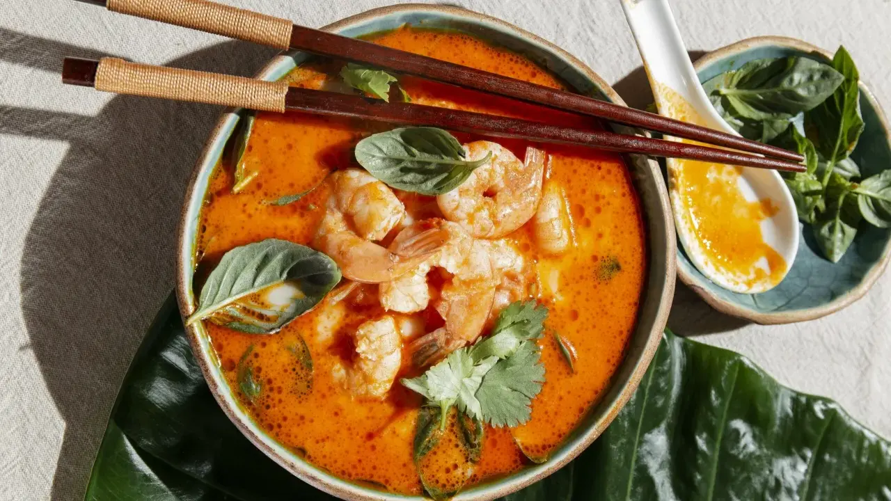 Miseczka zupy tom yum z krewetkami, ozdobiona ziołami, pałeczki i łyżka.