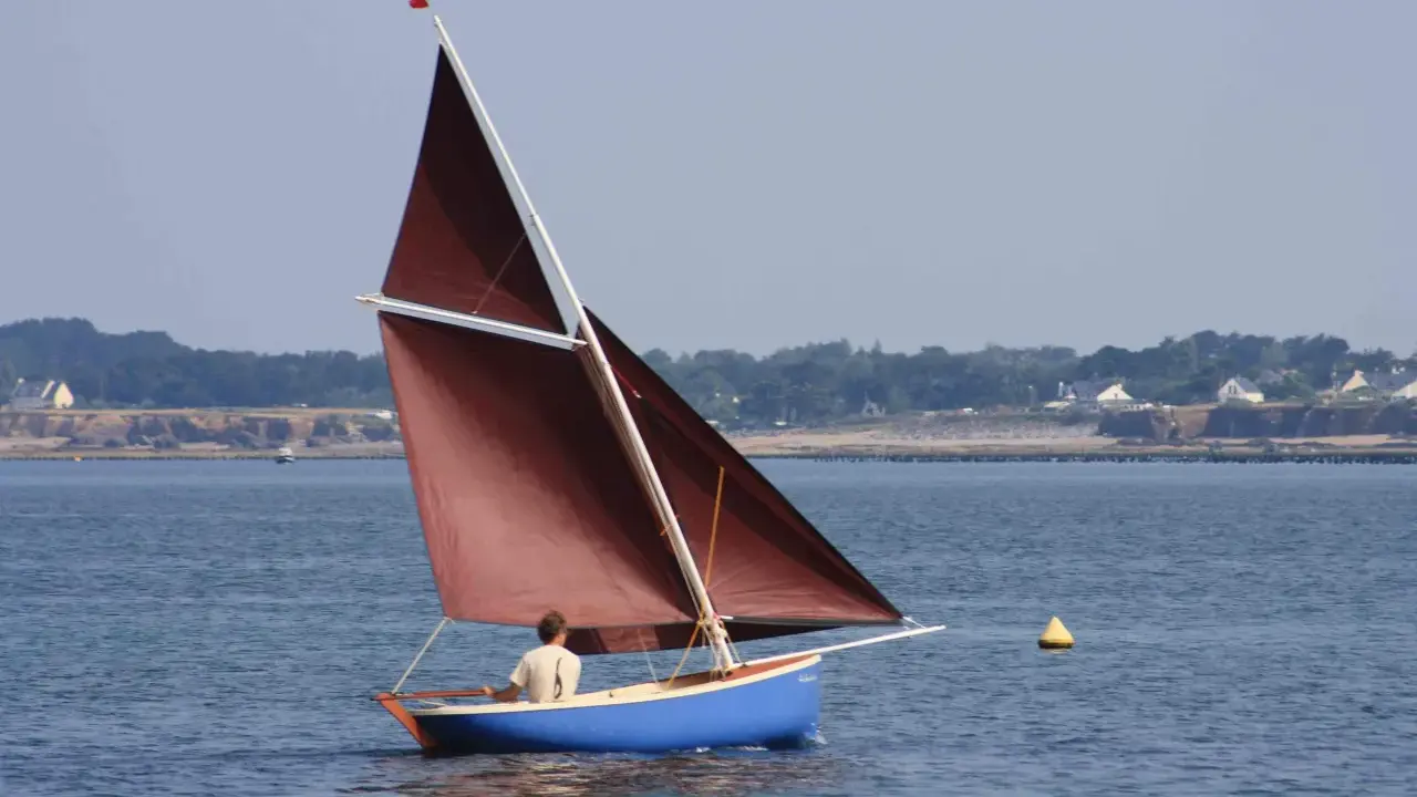 Un voilier bleu avec une voile marron navigue, tel une gazelle des îles, sur une mer calme. Au loin, une côte boisée bordée de maisons.
