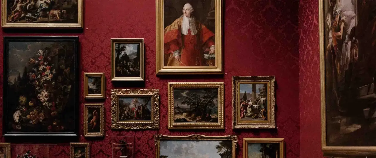 A gallery wall displays classical paintings in ornate frames against a rich red damask background. This collection showcases the best way to invest in art, with portraits and landscapes hinting at historical narratives.