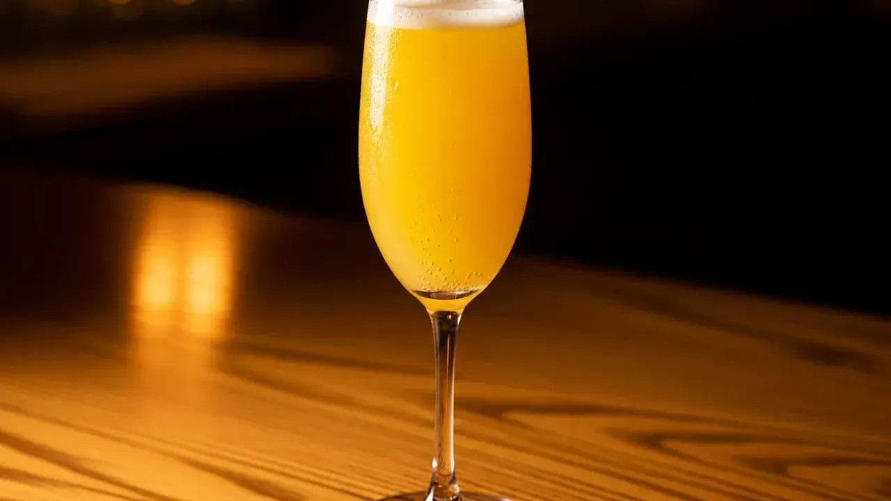 Ett glas med en gyllene mimosa drink, bubblig och uppfriskande, står på ett träbord i varmt ljus.