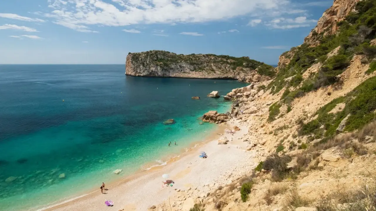 Türkisblaues Wasser trifft auf Kiesstrand in einer malerischen spanischen Atlantikbucht. Felsen und grüne Vegetation säumen die Küste.