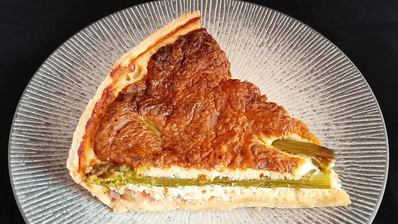 Tranche de tarte aux asperges blanches, garnie d'une préparation crémeuse et dorée, sur une assiette texturée.