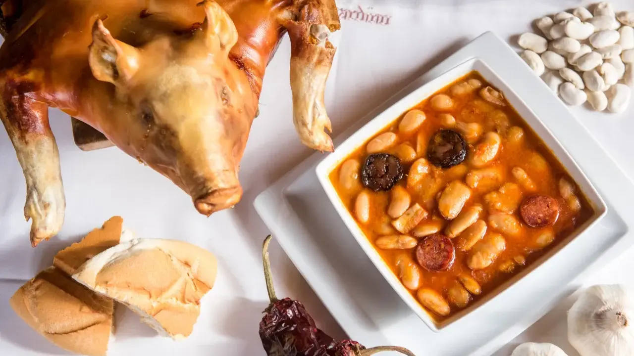 Cochinillo asado segoviano, crujiente y dorado, acompañado de un plato de judiones con chorizo y morcilla, pan y ajos.