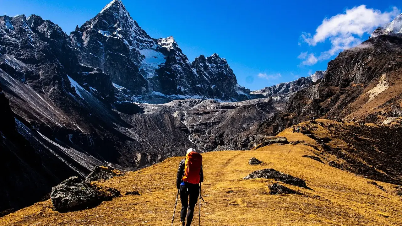 Osoba z plecakiem i kijkami idzie ścieżką przez górzysty, trawiasty teren. To jest właśnie trekking – podróżowanie pieszo po górach.