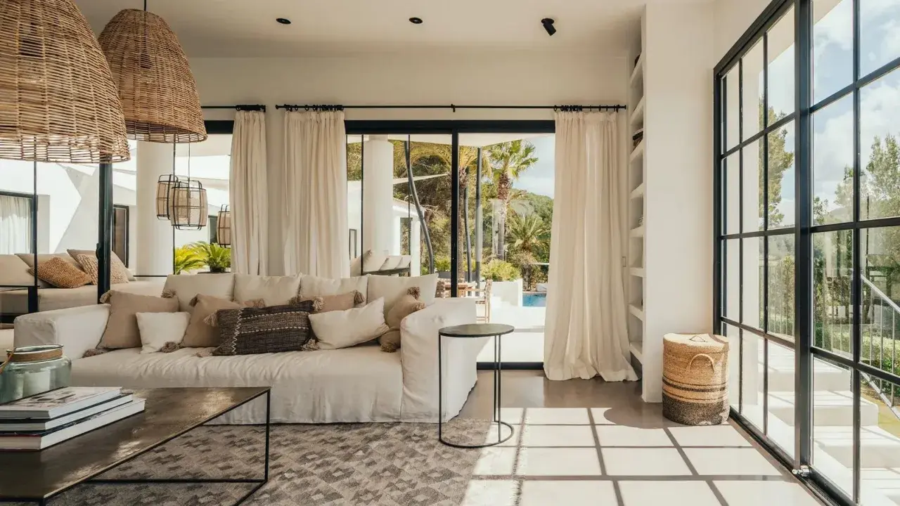 Arredare una casa al mare: divano bianco, cuscini naturali, lampade in rattan e vista su piscina e palme.