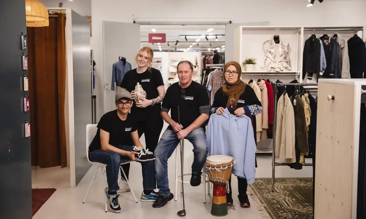 Fyra personer poserar i en butik med kläder och föremål. En kvinna håller en vas, en man en golfklubba, och en annan kvinna en ljusblå hoodie. De representerar hållbart second hand.