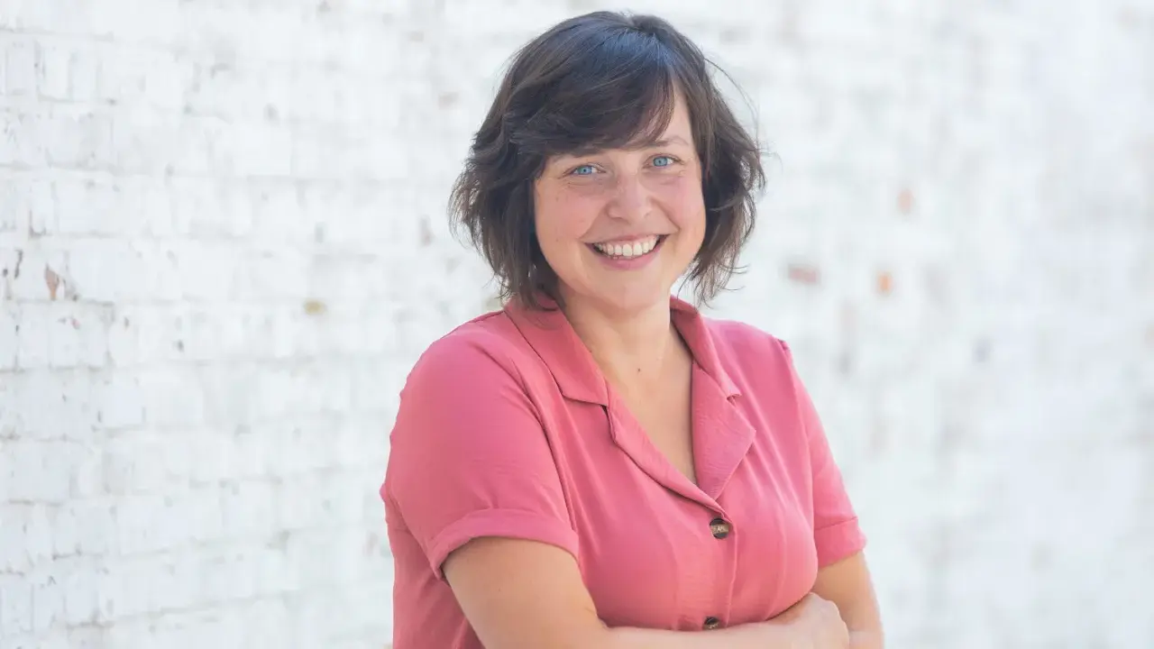 Een lachende vrouw met blauwe ogen, gekleed in een roze blouse, staat voor een witte bakstenen muur. Ze straalt de warmte uit van een zorgzame buurt.