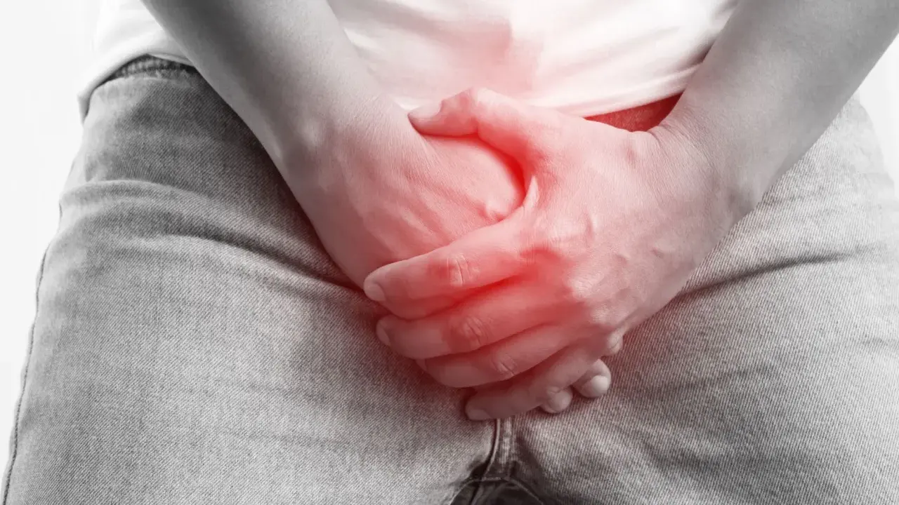 Dłoń obejmuje krocze z czerwonym podświetleniem, sugerując ból lub uraz, jak złamanie prącia.