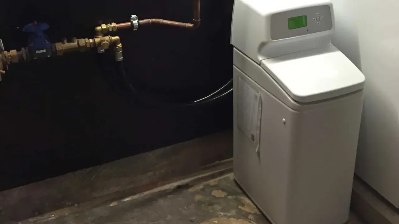 Appareil blanc avec écran vert, connecté à des tuyaux en cuivre. C'est un adoucisseur d'eau potable pour une maison.
