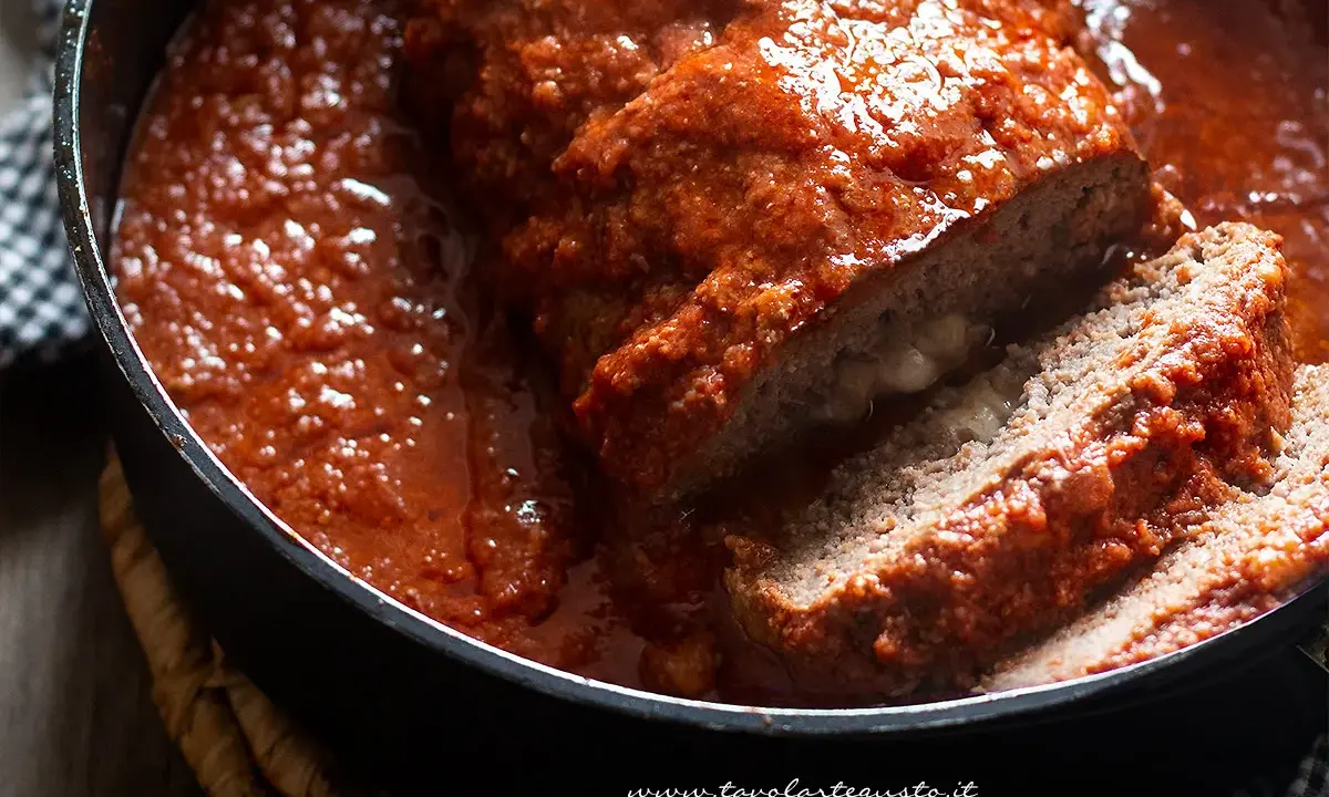 Un succulento polpettone al forno, affogato in un sugo rosso intenso, con una fetta tagliata che rivela un cuore filante di formaggio.