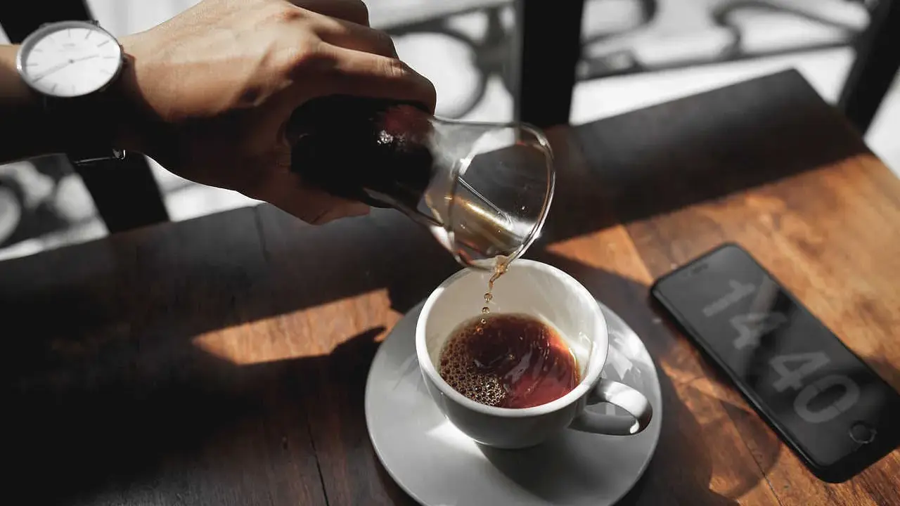 Hand gießt Kaffee in eine Tasse, daneben ein Smartphone mit Uhrzeit.