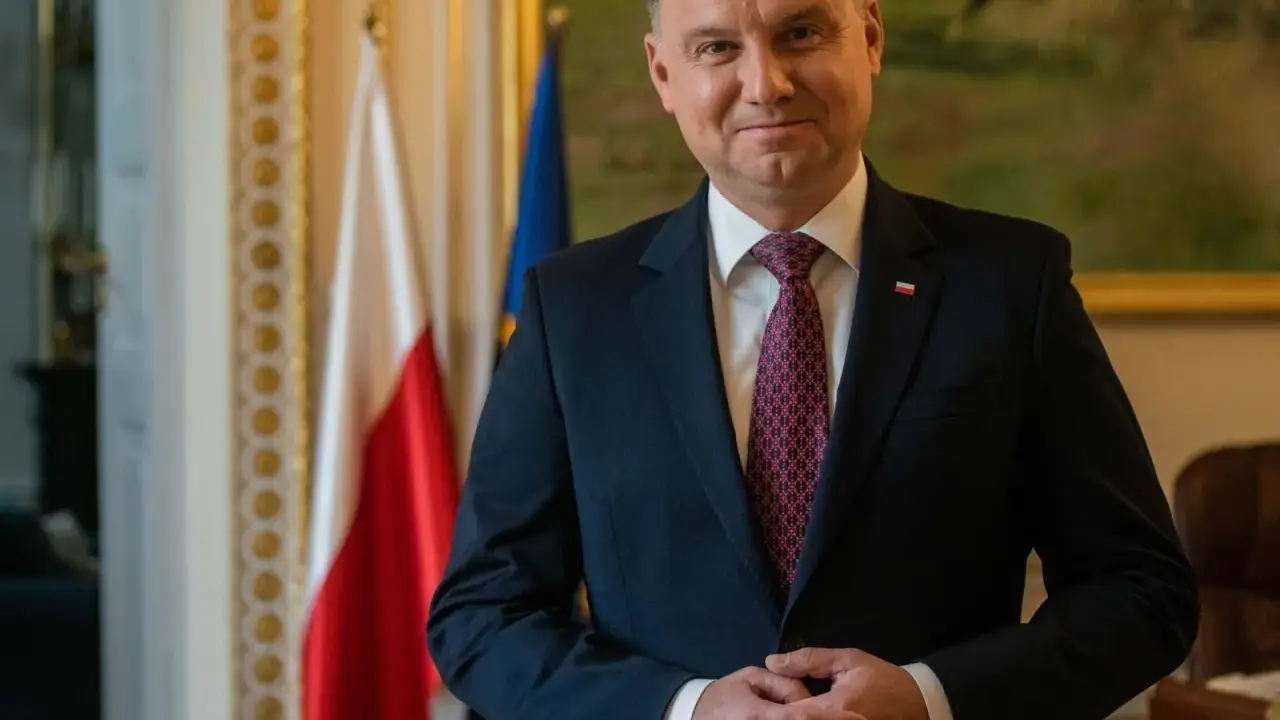 Prezydent Andrzej Duda w garniturze, z polską flagą w tle, czeka na moment, kiedy prezydent podpisze emerytury stażowe.