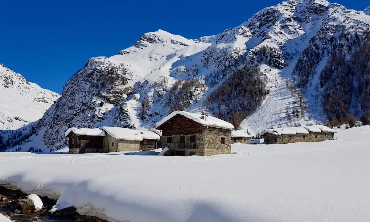 Case innevate nel Val di Rezzalo, con baite in pietra e legno. Per sapere val di rezzalo come arrivare, segui il ruscello che scorre tra la neve.