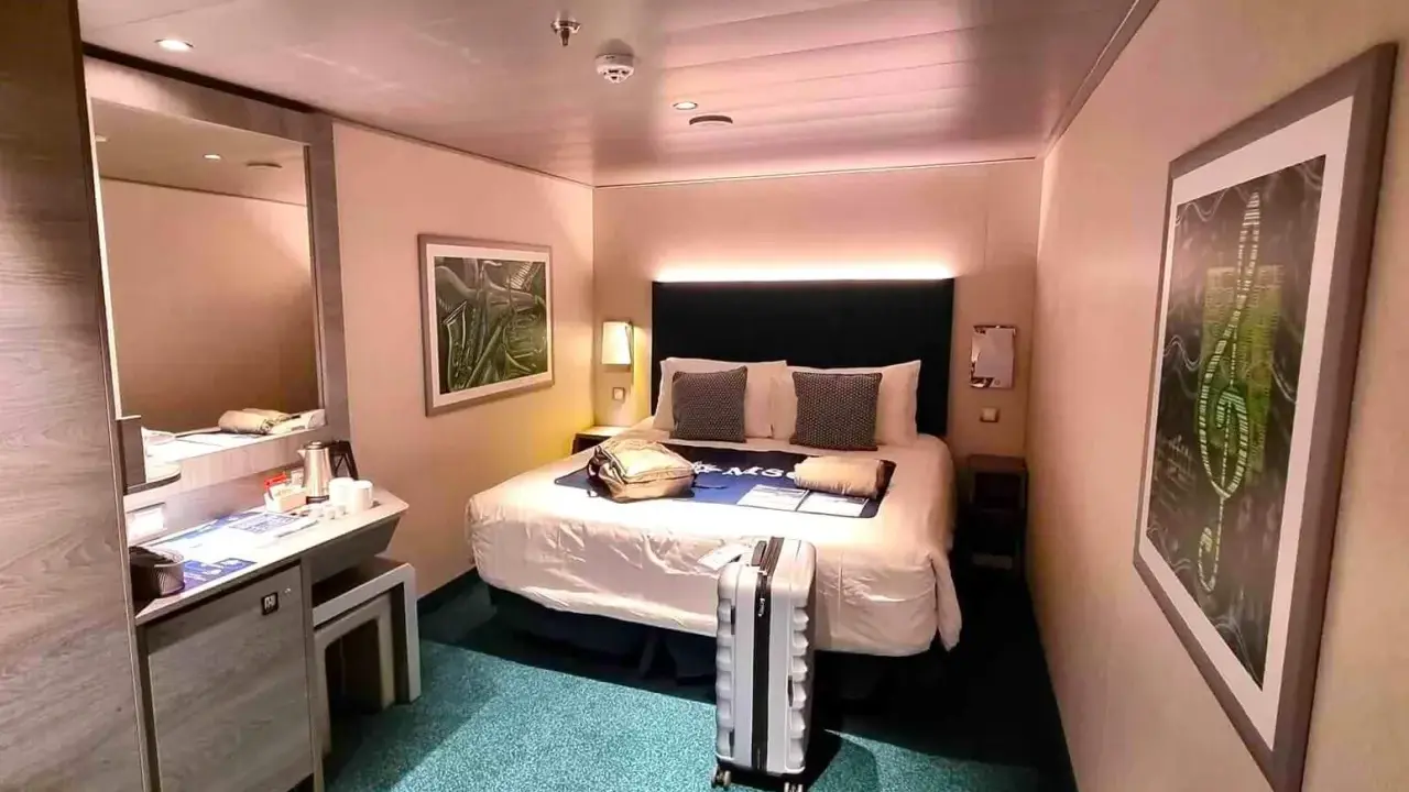 Interno di una royal caribbean cabine con letto matrimoniale, specchio, scrivania e valigia pronta per il viaggio.