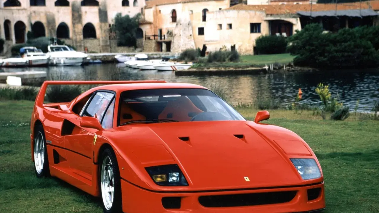 Roter Ferrari F40 auf einer Wiese, im Hintergrund ein Hafen mit Booten und Häusern. Der Ferrari F40 Neupreis war damals ein Traum.