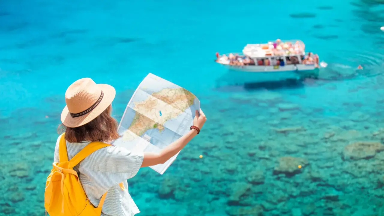 Ragazza con zaino giallo e cappello di paglia studia una mappa, pensando a quanti soldi portare in crociera, con mare turchese e barca sullo sfondo.