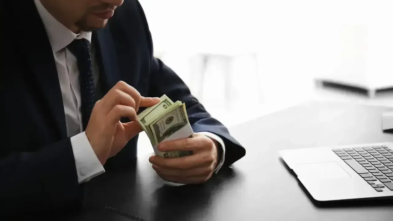 Mężczyzna w garniturze liczy pieniądze, zastanawiając się, ile zarabia osoba pracująca w banku. Obok leży laptop.