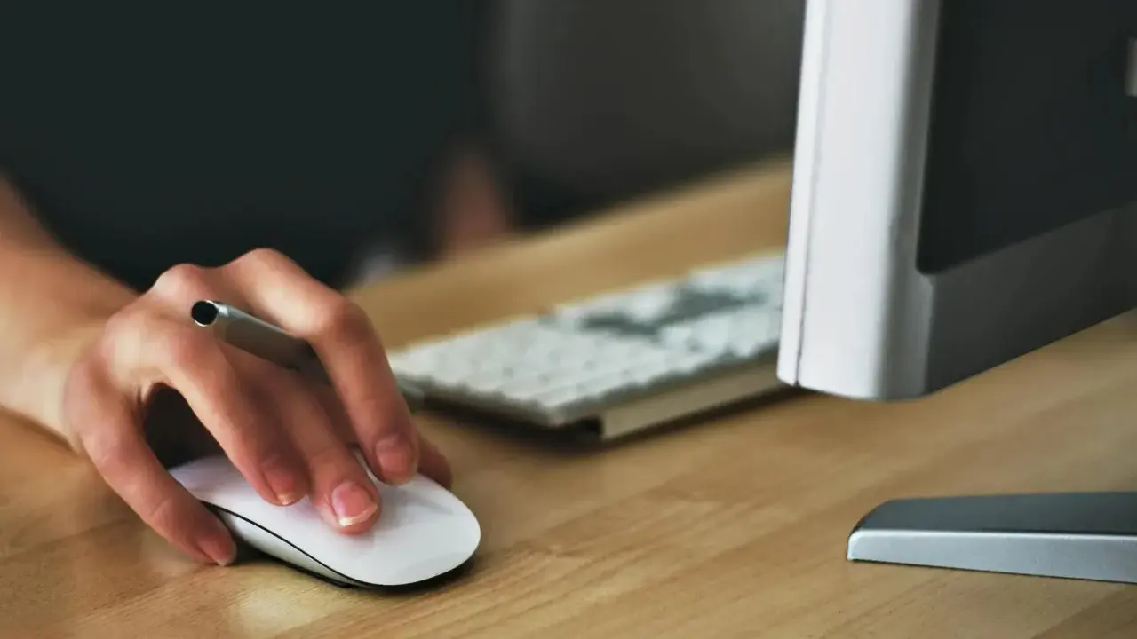 Main souris blanche sur un bureau en bois, avec un clavier et un écran d'ordinateur flous en arrière-plan.