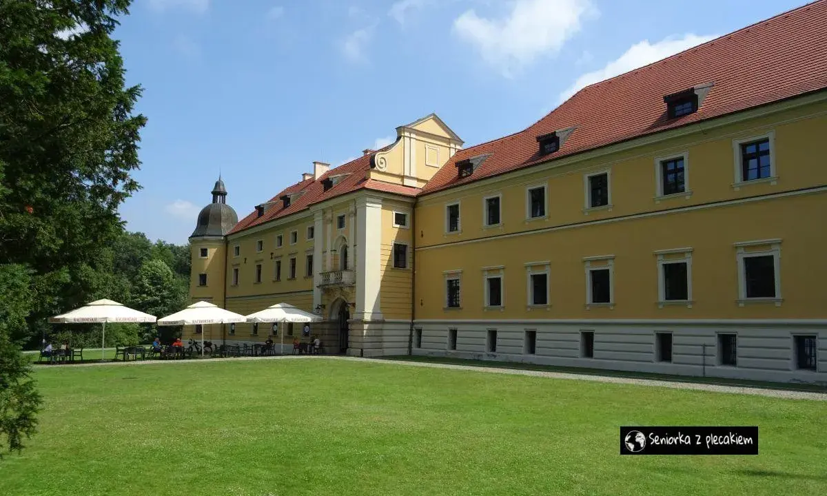 Żółty pałac z czerwonym dachem, gdzie można usiąść pod parasolami. W oddali widać drzewa, a na trawniku rozłożono stoliki. To miejsce, gdzie można poczuć atmosferę rudy raciborskie.