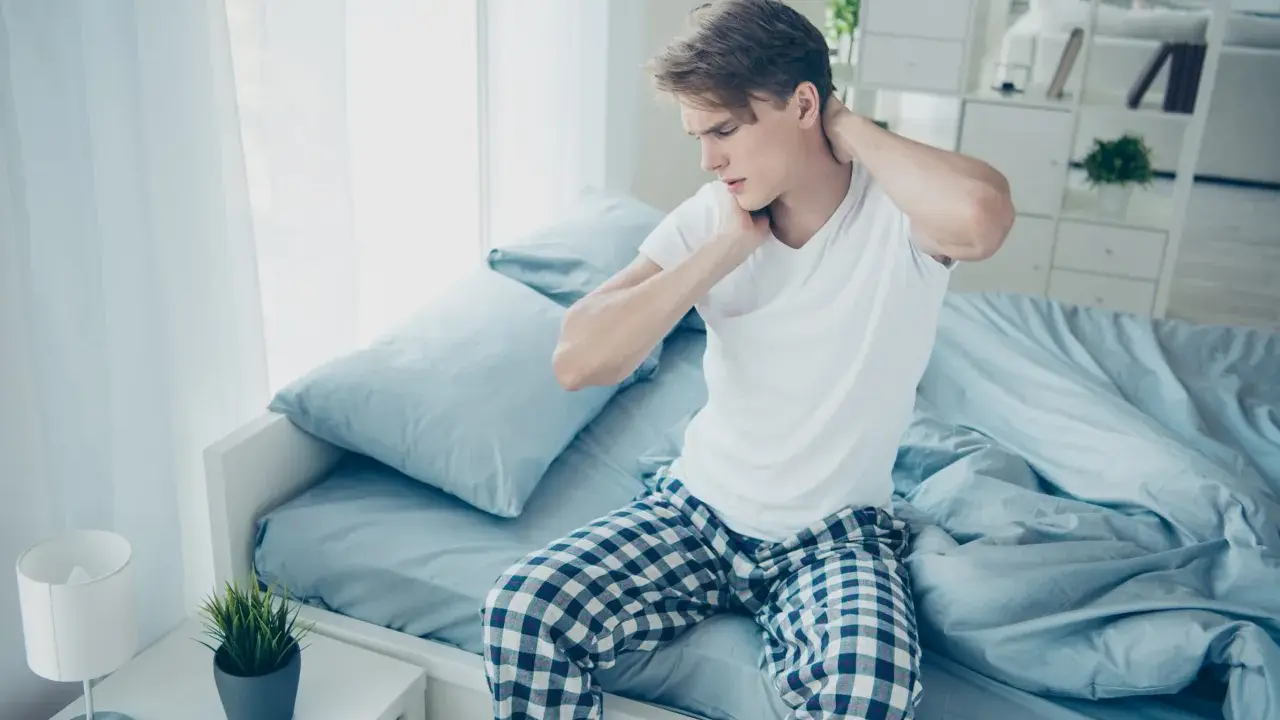 Junger Mann sitzt auf dem Bett, reibt sich den Nacken. Er hat nackenschmerzen nach dem schlafen.