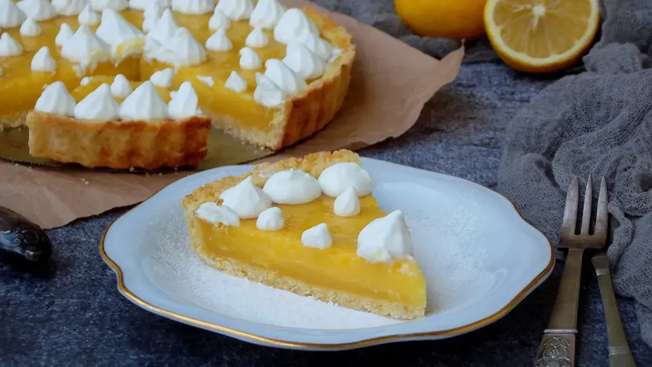 Une part de tarte au citron, garnie de meringue, sur une assiette. La tarte entière est visible en arrière-plan. Une recette de tarte au citron parfaite pour le dessert.