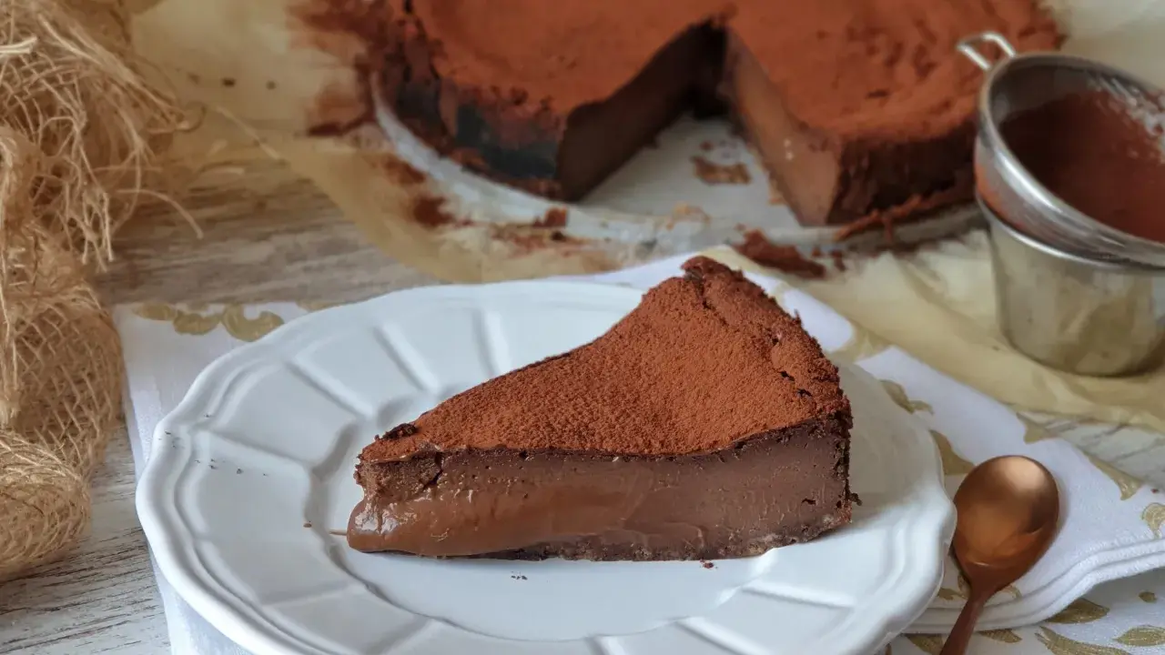 Una porción de tarta de chocolate, cubierta de cacao en polvo, reposa en un plato blanco. El resto de la tarta, también espolvoreada, se ve al fondo.
