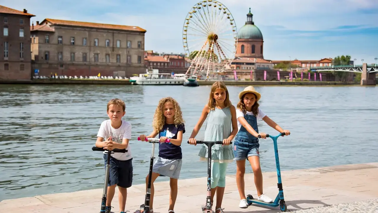 Niños en patinetes junto al río en Toulouse, con la noria y la cúpula de la Basílica de San Sernín de fondo. ¡Un plan genial que hacer en Toulouse con niños!