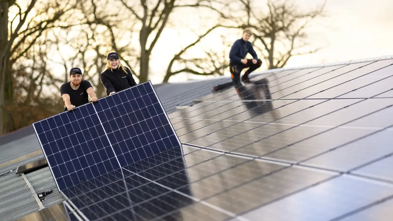 Arbetare installerar solpaneler på ett tak. En av dem, en kvinna, ler glatt medan hon håller i en panel. En annan man hjälper till, och en tredje person sitter på taket. De är en del av teamet för **solverkets alfa**.