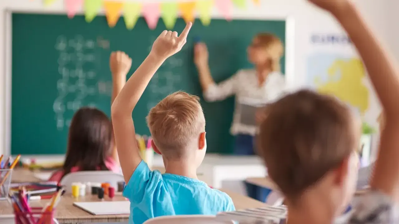 Uczniowie z podniesionymi rękami na lekcji. Nauczycielka pisze na tablicy. To obrazek, który mógłby znaleźć się na karcie nauczyciela.