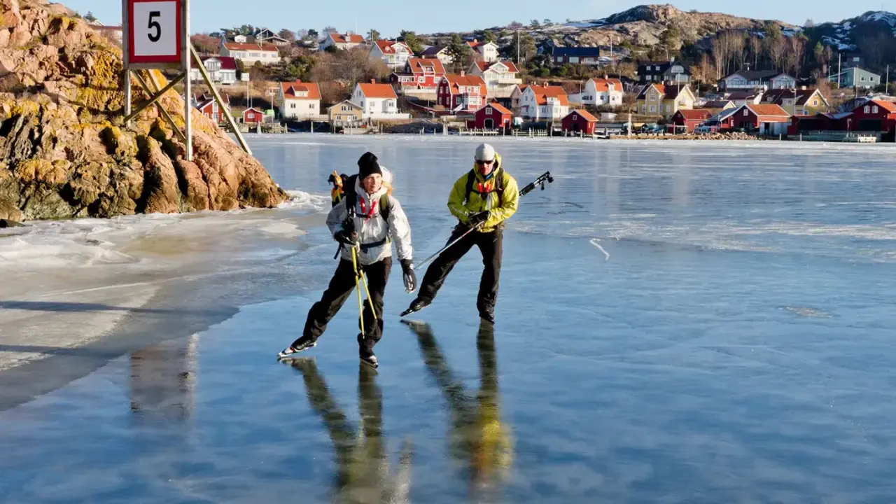 Två personer åker skridskor på en isbelagd sjö, med en pittoresk by i bakgrunden. De använder stavar för att hjälpa sig fram, vilket visar hur åker man skridskor på ett effektivt sätt.