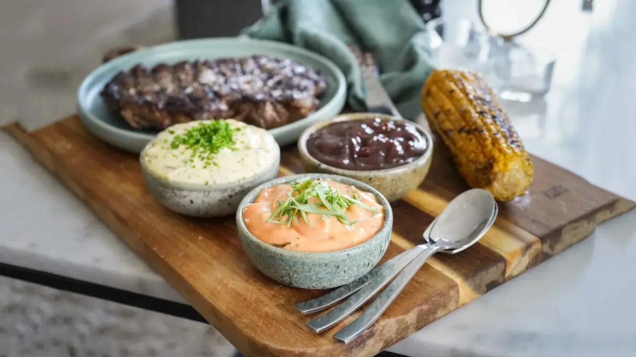 Grillad biff med tre olika såser till grillat: en vit med örter, en mörk och en orange. Majskolv och bestick ligger på en träbricka.
