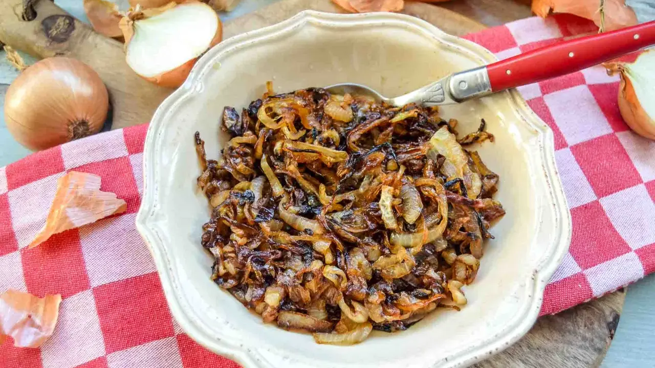 Schüssel mit karamellisierten Zwiebeln, die auf einem karierten Tuch liegen. Ganze und halbierte Zwiebeln sind daneben zu sehen.