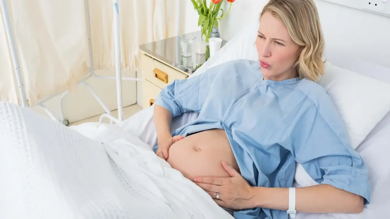 Gravid kvinna i sjukhussäng, med händerna på magen, ser ut att ha värkar. Vattnet har gått och hon väntar på barnet.