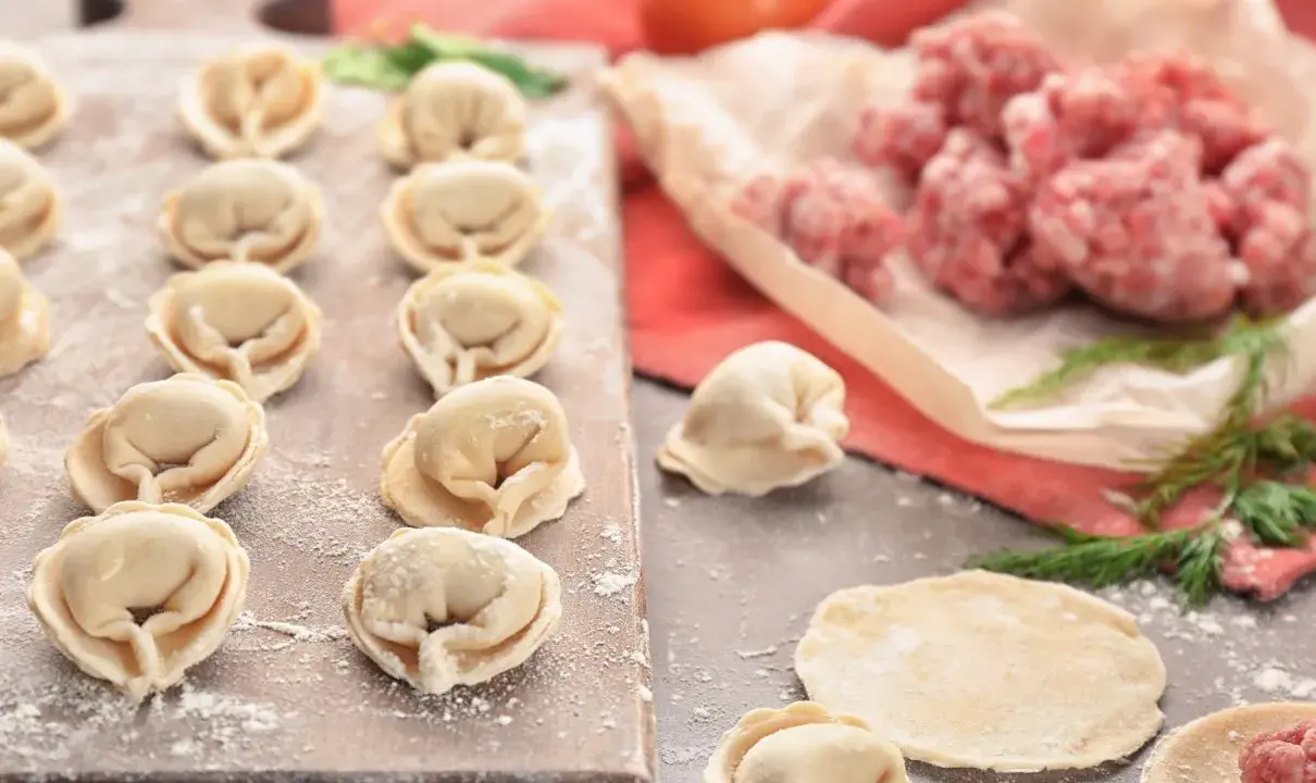 Gotowe do lepienia pierogi z mięsem mielonym surowym. Na desce ułożone pierożki, obok surowe mięso i ciasto.