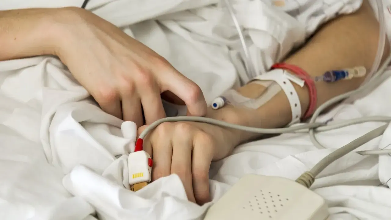 Ręka pacjenta w szpitalnym łóżku, podłączona do aparatury medycznej. Denga to choroba, a tu widzimy jej skutki.