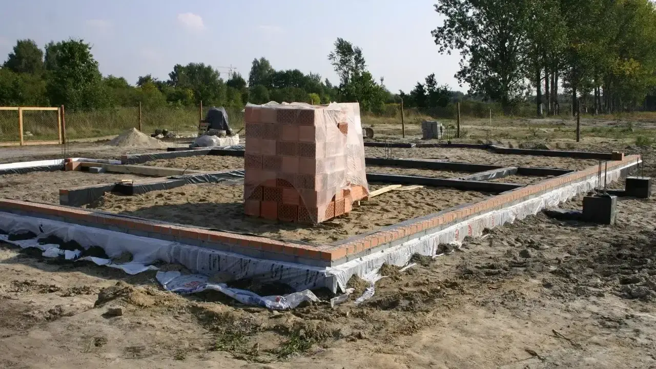 Stan zero budynku: fundamenty z cegły, piasek, folia i stos cegieł pod przykryciem.