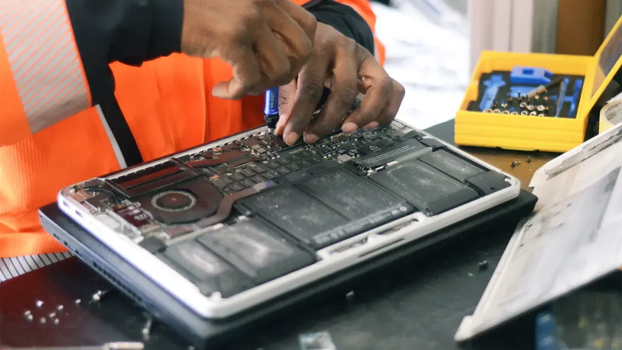 Réparation d'un ordinateur portable ouvert, mains expertes utilisant un tournevis. Une boîte à outils jaune est visible, prête pour la prochaine étape, sans deee taxe.