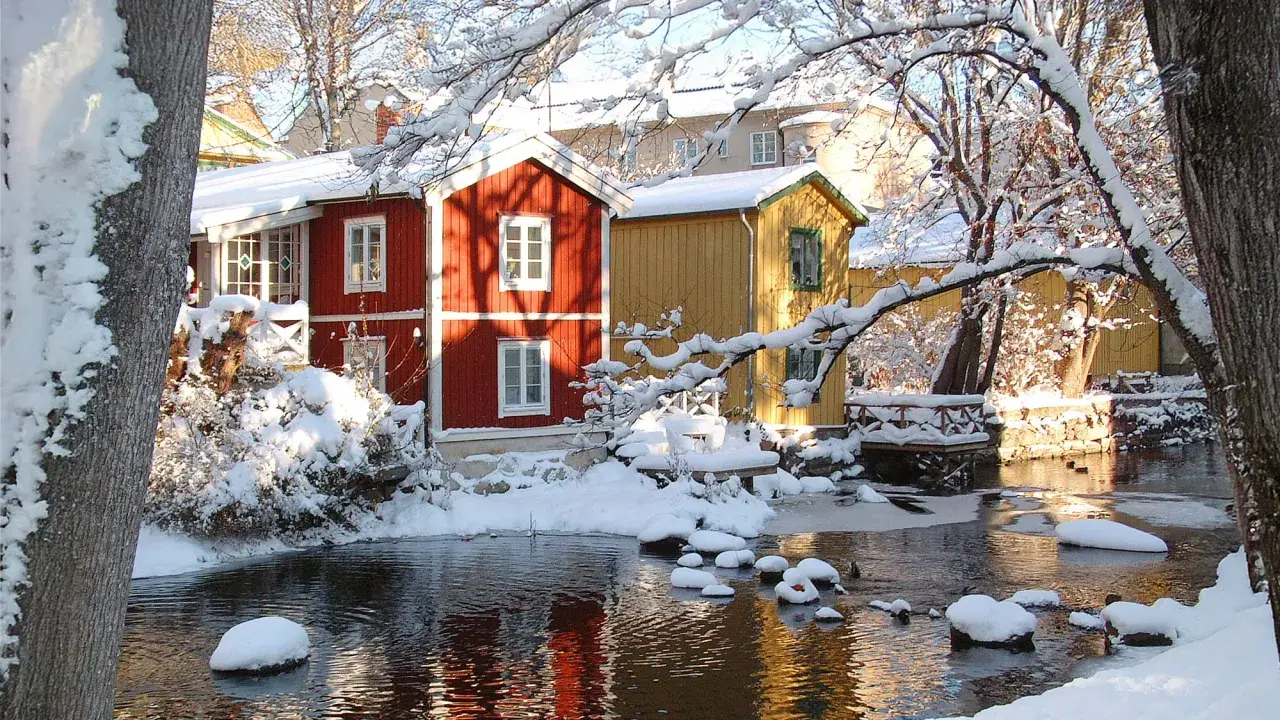 Vinteridyll vid vattnet i destination Gåsvik. Röda och gula hus speglas i det isiga vattnet, omgivna av snötyngda träd.