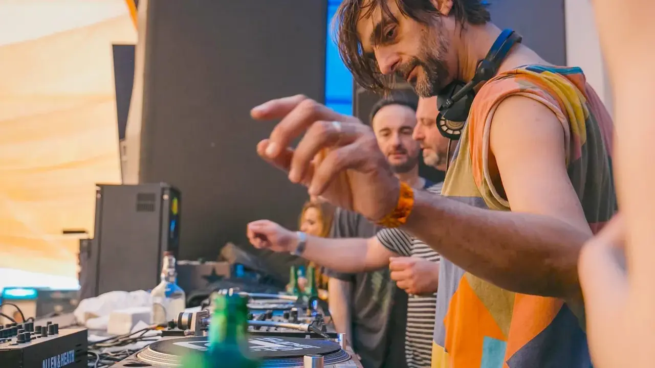 DJ Villa Lobos, con cuffie al collo, mixa musica con passione. Dietro di lui, il pubblico attende il ritmo.