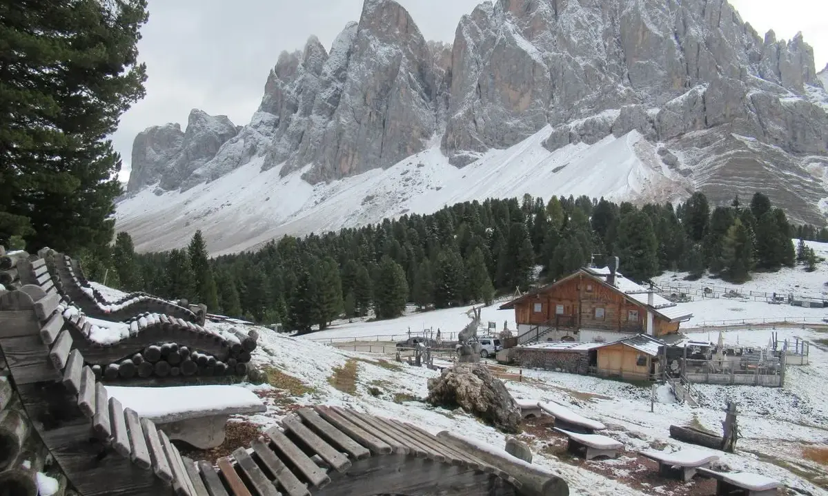 Panorami innevati con il rifugio delle Odle in lontananza, panchine in legno e pini.