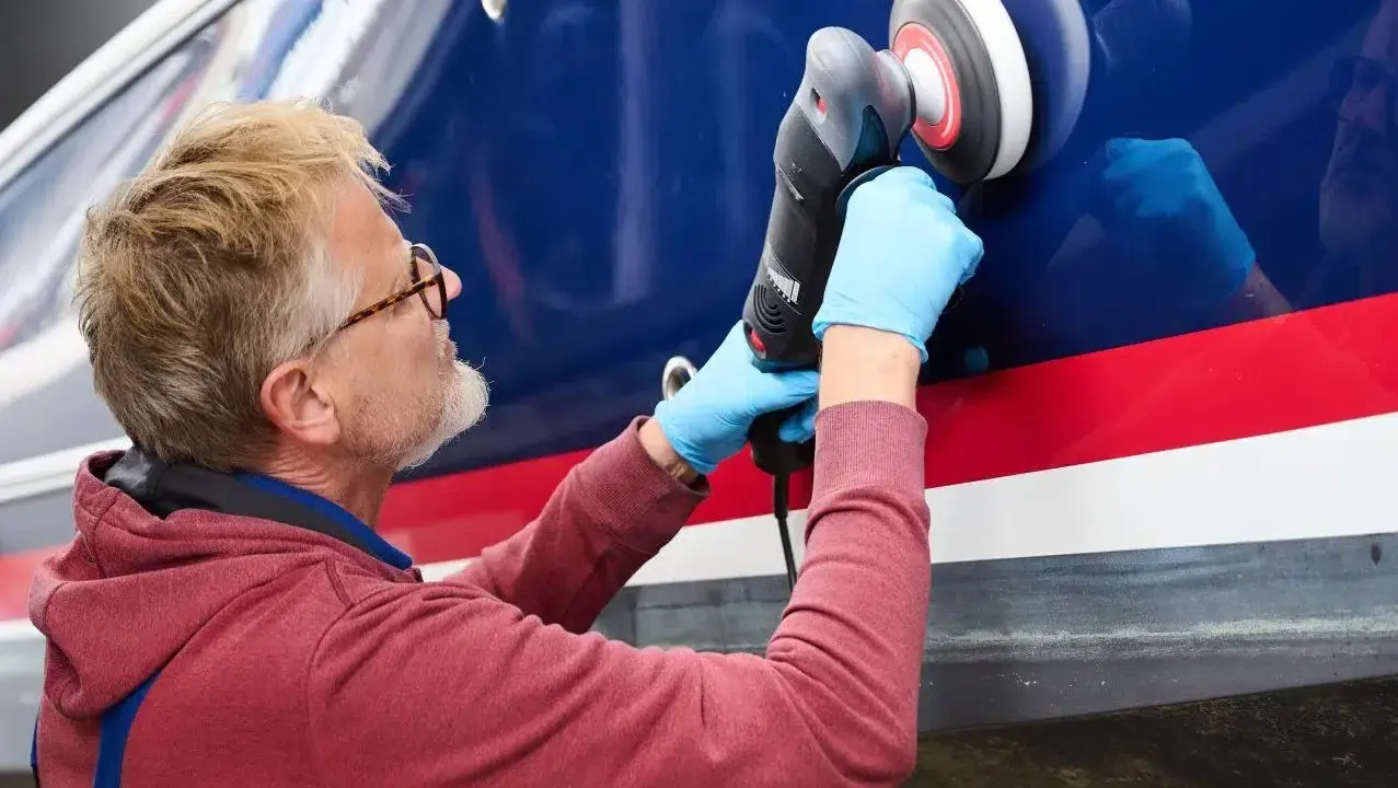 Mann poliert mit Maschine den blauen Bootsrumpf. Das Gelcoat erstrahlt nach dem Polieren wieder in neuem Glanz.