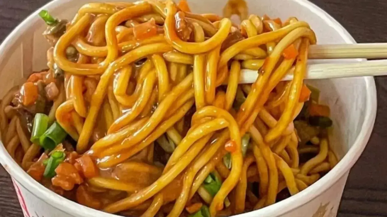 Un tazón lleno de fideos chinos salteados con verduras y salsa. Palillos levantan un bocado.