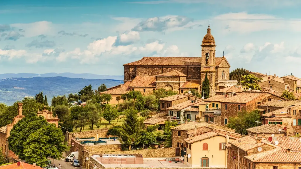 Panoramica del centro storico di Montalcino, con la chiesa e le case in pietra sotto un cielo azzurro con nuvole.