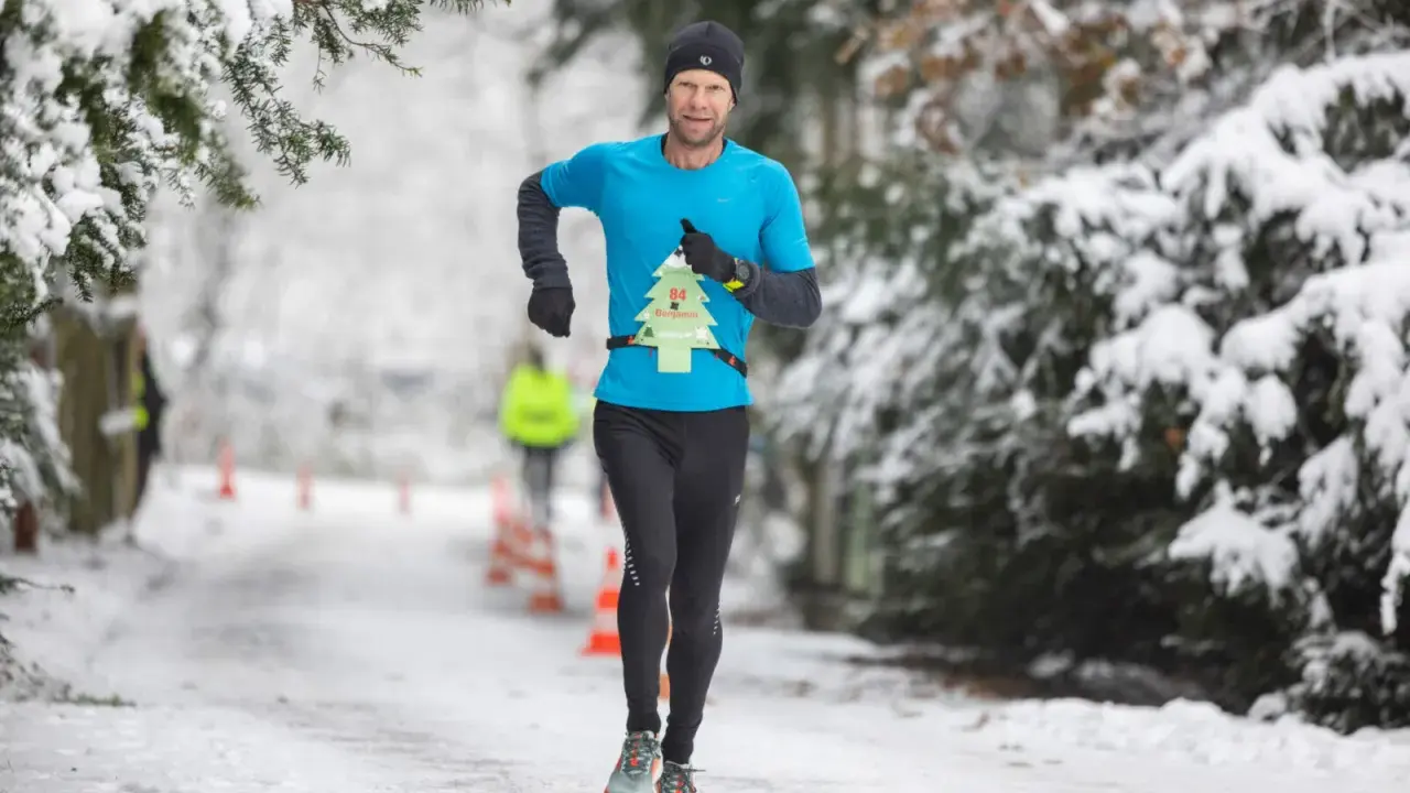 Mann läuft im Winter durch verschneiten Park. Er trägt ein blaues Shirt mit einer Startnummer in Form eines Weihnachtsbaums.