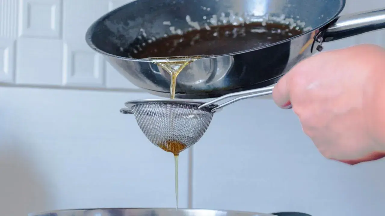 Se cuela aceite rancio de una sartén a través de un colador fino, cayendo en un recipiente.