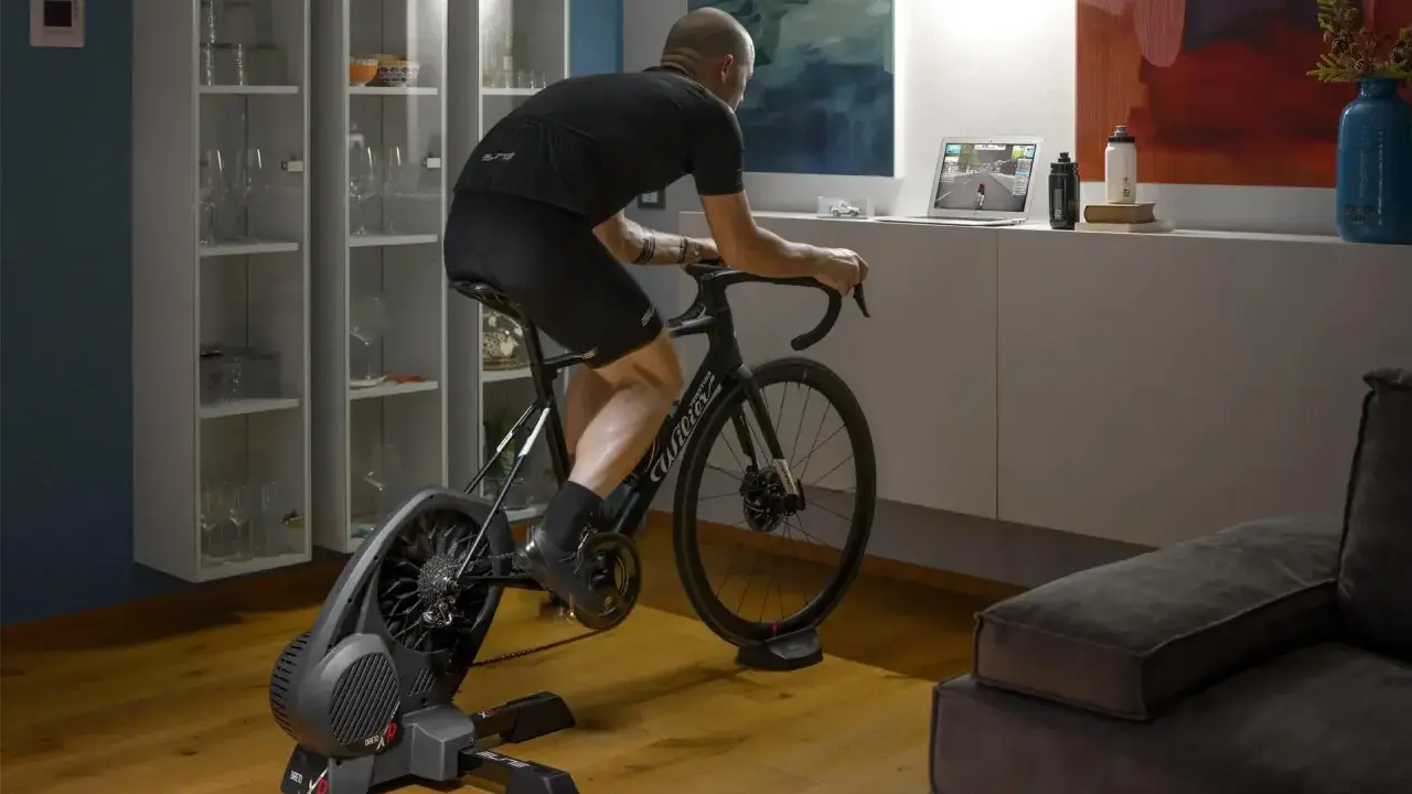 Mężczyzna trenuje na rowerze stacjonarnym z napędem **direct drive**, śledząc wirtualną trasę na laptopie.