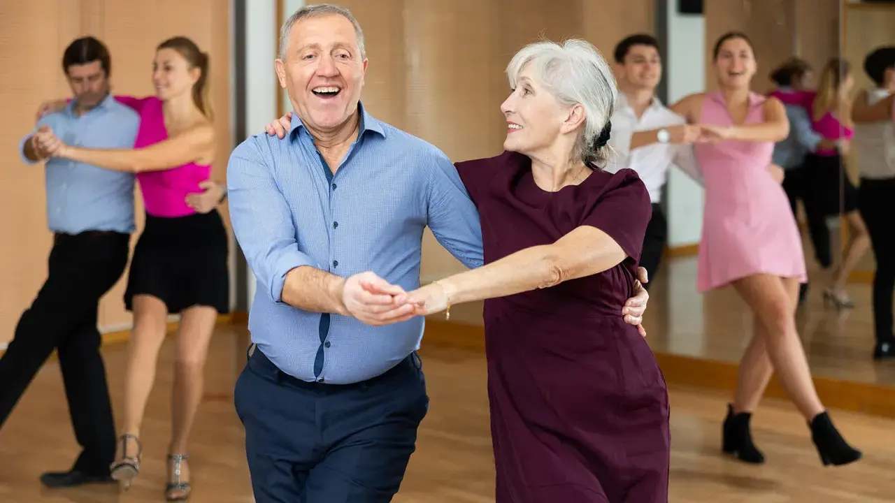 Uśmiechnięta para seniorów uczy się, jak tańczyć na 3, w tle inne pary.