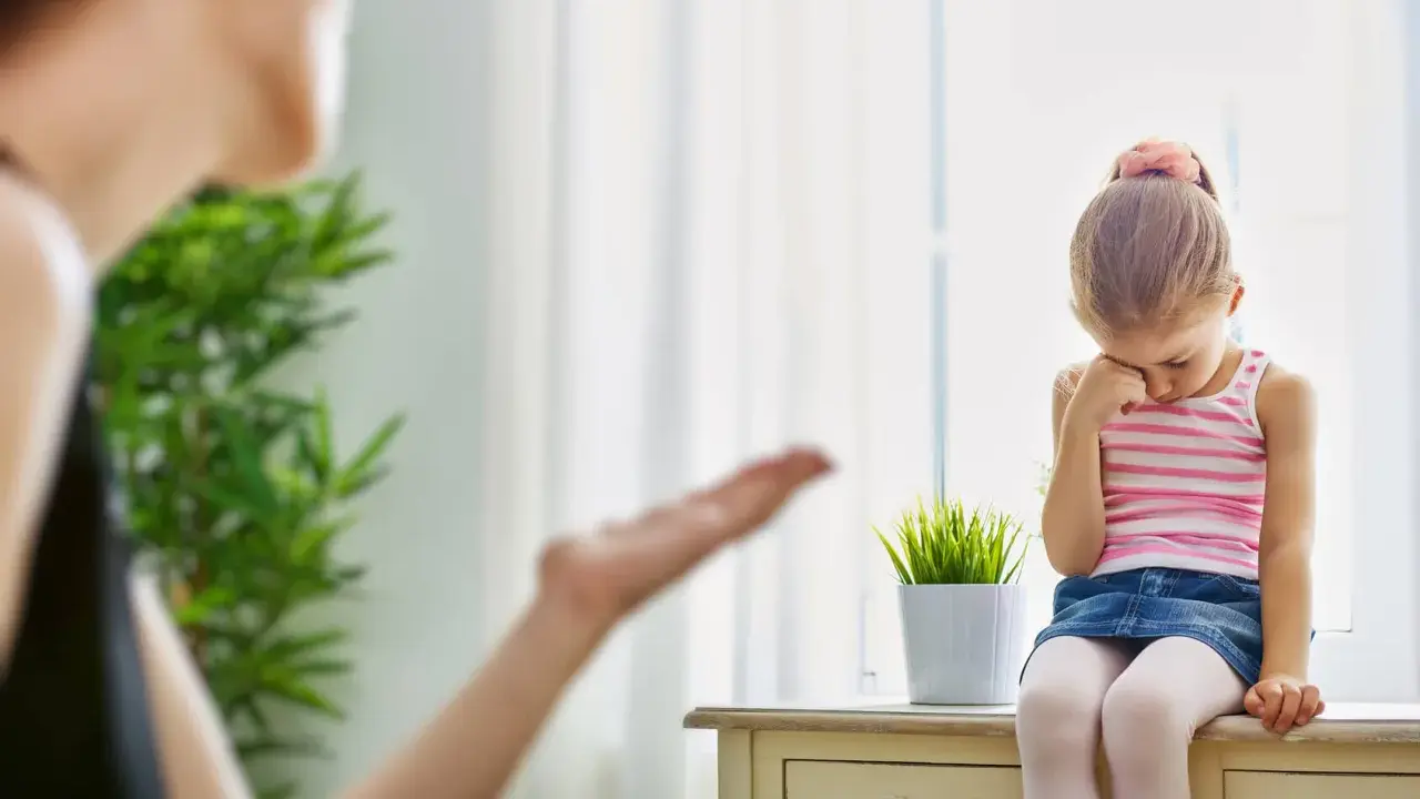 Smutna dziewczynka siedzi przy oknie, a jej mama gestykuluje. Warto nauczyć się, jak przestać krzyczeć na dziecko, by budować lepsze relacje.