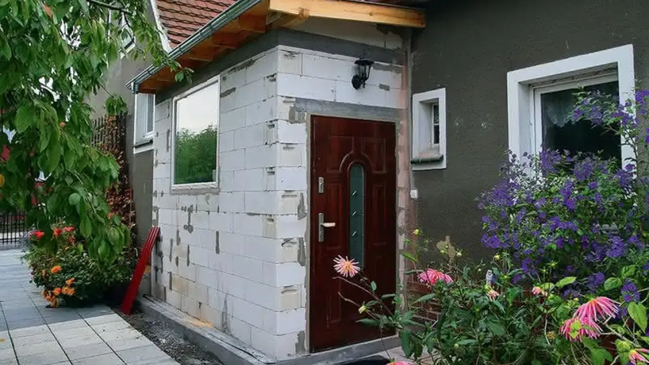 Budowa dobudowy wiatrołapu z bloczków, z nowymi oknami i drzwiami, otoczona zielenią.